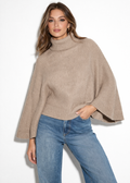Woman wearing a BCBG Women's Oversized Turtleneck Sweater Poncho and blue jeans on a white background